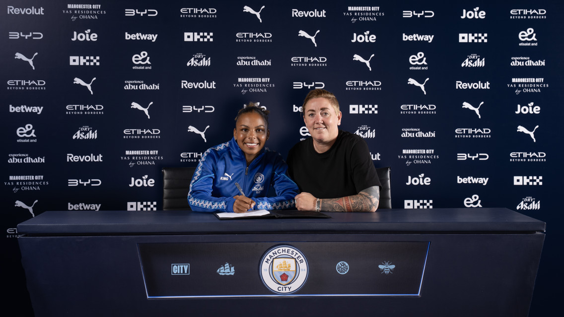 Two people sitting at a desk with Manchester City logos and sponsor background. One is signing a document.